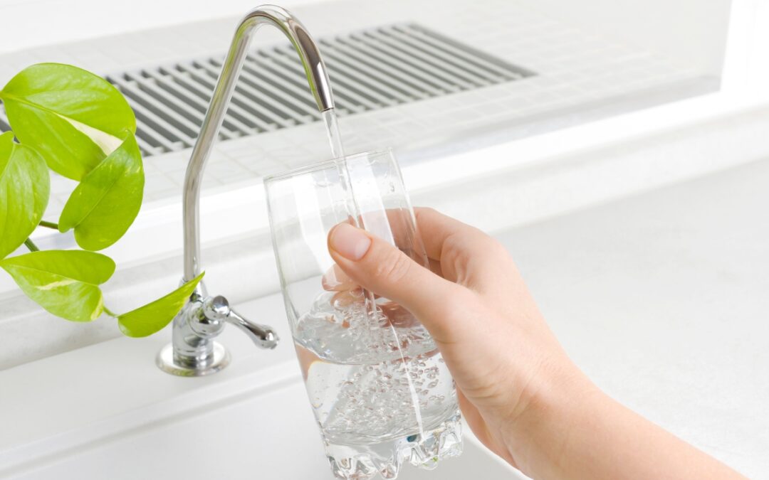 A hand holds a glass under a faucet, filling it with cold, filtered water in a bright kitchen setting. A green plant is visible next to the faucet.