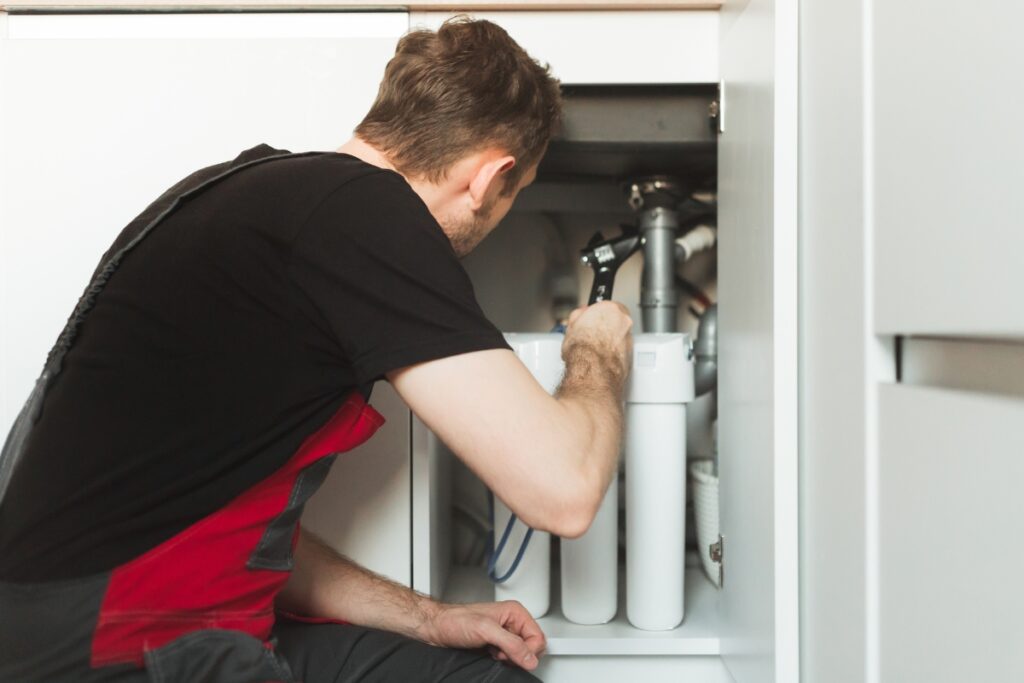 A person works under a sink, focusing on the plumbing or filter system in a corporate office refreshment area, wearing a black and red outfit.