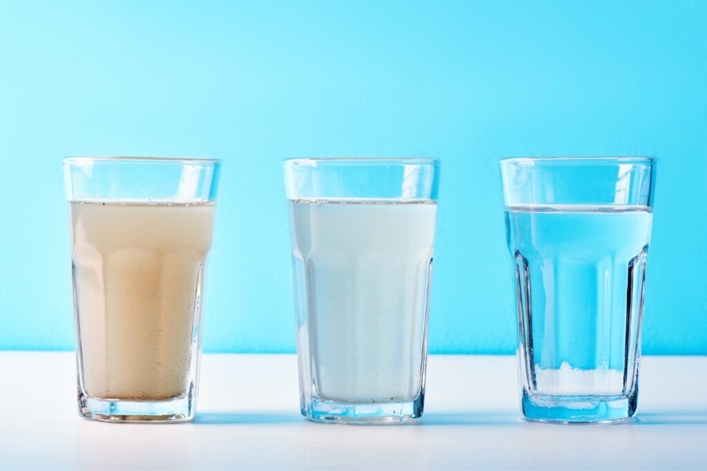 Three glasses of water, resembling paper products in their simplicity, are placed side by side on a white surface against a blue background. The water appears cloudy, partially clear, and clear respectively.