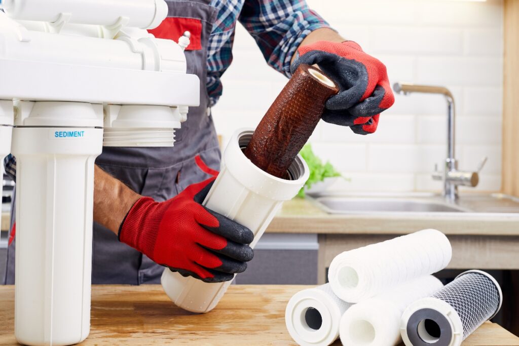 A person wearing red gloves replaces a used filter in a water filtration system from Coffee Man Beverage Services while clean filters lie on the table.
